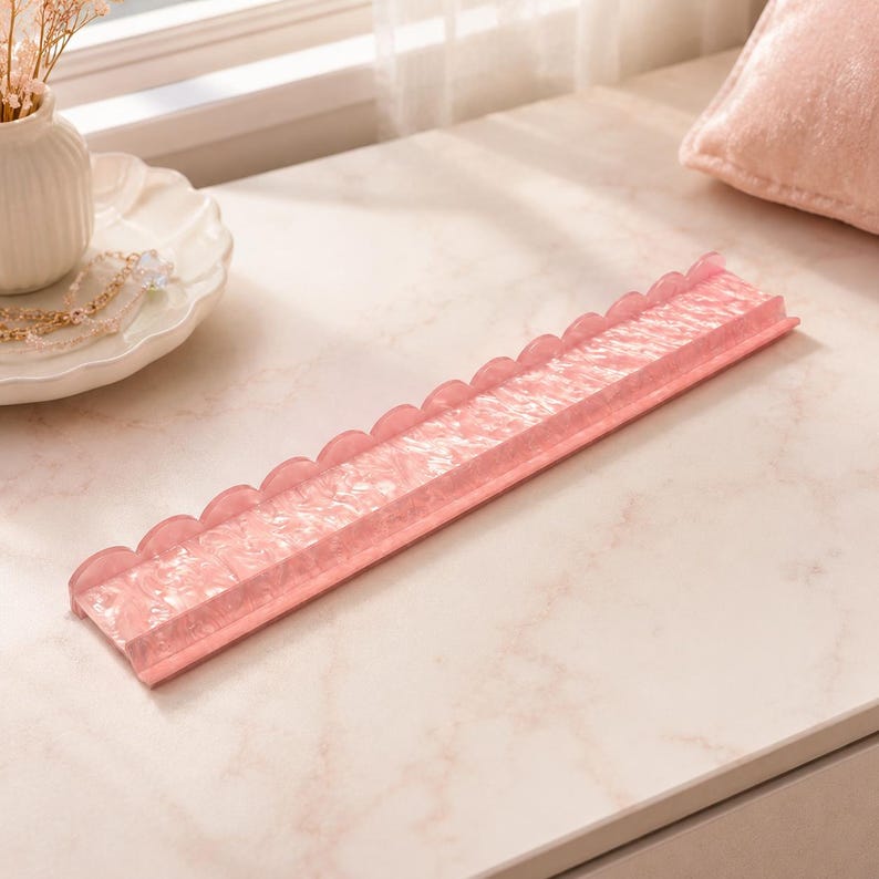 May include: A pink, scalloped-edge jewelry tray. The tray is rectangular with a textured surface and a raised edge. The tray is made of a translucent material and sits on a light-colored surface. The tray is designed to hold jewelry.