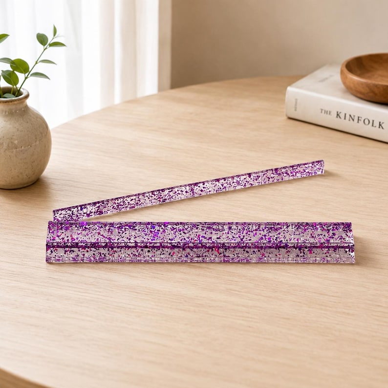 May include: Two clear, rectangular purple glitter rulers are displayed on a light wood surface. One ruler is lying flat, while the other is angled upwards. The glitter is a mix of purple and silver.