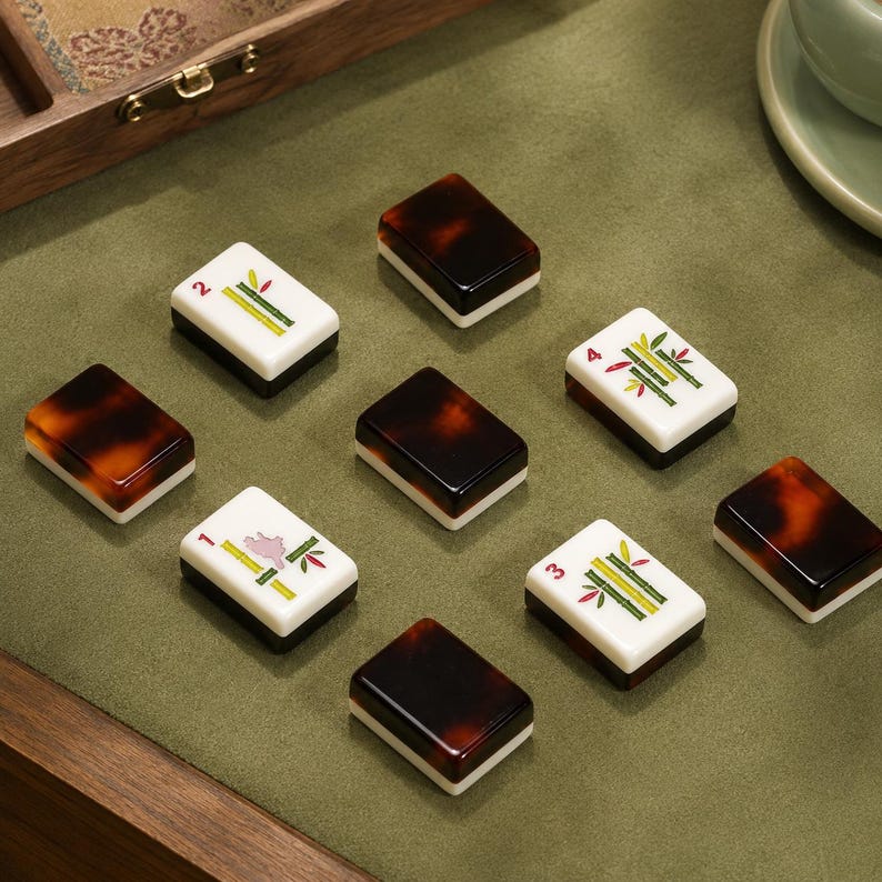 May include: A set of rectangular mahjong tiles featuring a two-toned design with a dark brown tortoiseshell pattern on one side and a white face on the other. Some tiles display green bamboo illustrations with numbers one through four in red.