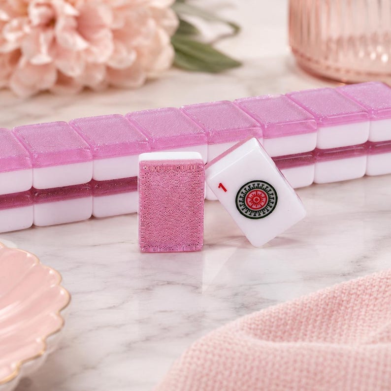 May include: A set of pink and white Mahjong tiles. One tile is white with a red and black design and the number 1. Another tile is pink with a textured surface. The tiles are arranged on a marble surface.