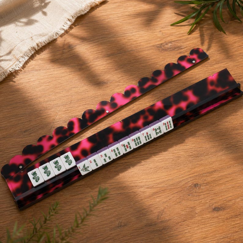 May include: A pink and black Mahjong tile rack with a matching tile holder. The rack and holder have a tortoiseshell pattern. The tiles are white with green and red symbols. The set is on a wooden surface.