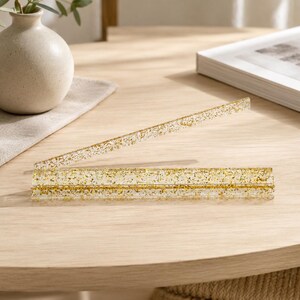 May include: Clear acrylic photo holder with gold glitter embedded throughout. The holder is rectangular with a slot to hold a photo or card. A matching clear acrylic stick is also included.
