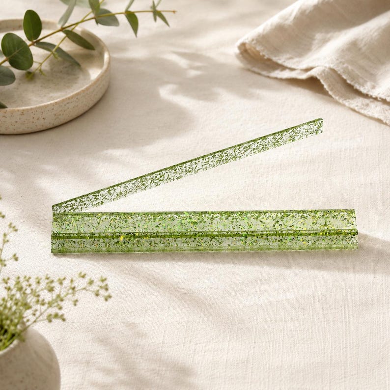 May include: A clear, green, glitter-infused acrylic letter opener. The letter opener is open, with one section angled upward. The background includes a white plate, a sprig of greenery, and a beige cloth.