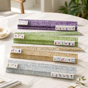 May include: Four colorful Mahjong tile racks in purple, green, gold, and silver, each holding a set of tiles. The racks have a glittery, textured appearance, and the tiles display various symbols and characters. The scene is set on a white table.