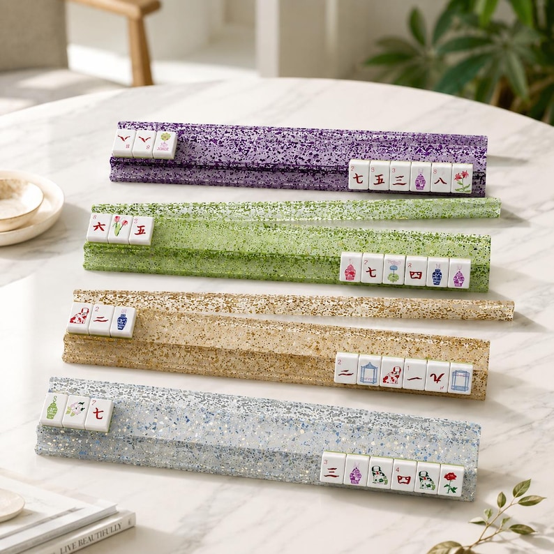 May include: Four colorful Mahjong tile racks in purple, green, gold, and silver, each holding a set of tiles. The racks have a glittery, textured appearance, and the tiles display various symbols and characters. The scene is set on a white table.
