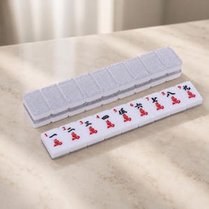 May include: A white Mahjong tile set with red symbols and numbers. The tiles are arranged next to a silver glitter case. The set is on a light-colored surface.