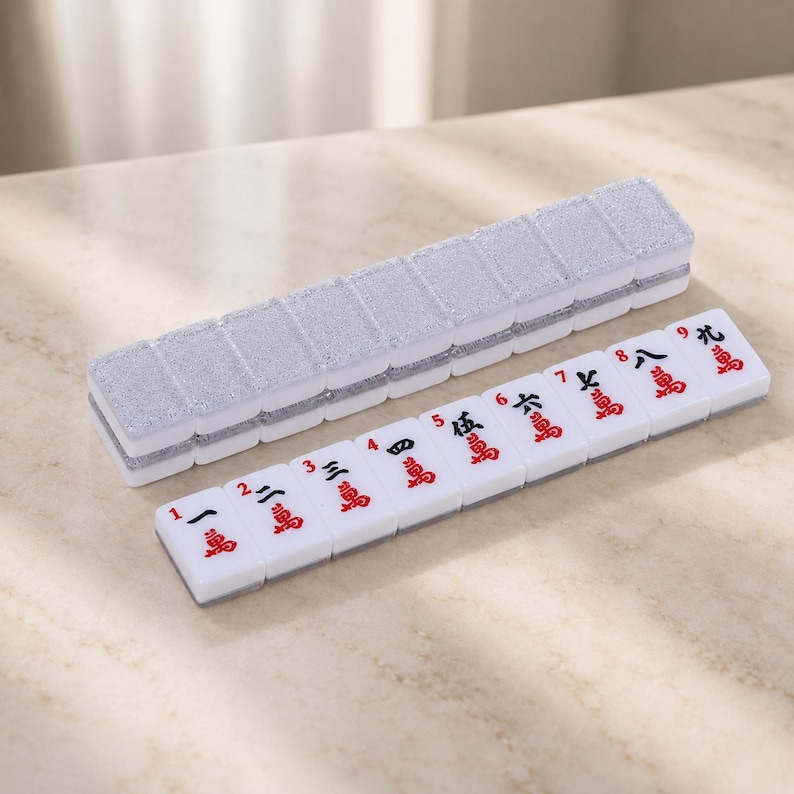 May include: A white Mahjong tile set with red symbols and numbers. The tiles are arranged next to a silver glitter case. The set is on a light-colored surface.