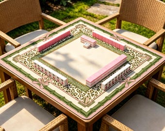 May include: A square wooden mahjong table with a cream-colored playing surface, surrounded by pink and green decorative borders. Pink and white mahjong tiles are arranged on the table. Four wicker chairs surround the table, set on a grassy lawn.