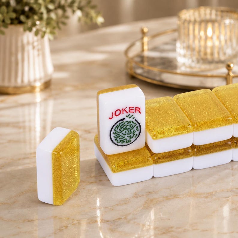 May include: White and gold Mahjong tiles with the word "JOKER" and a green and black design. The tiles are arranged on a marble surface, with a candle holder and decorative plant in the background.