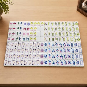 May include: A complete set of white mahjong tiles arranged in a grid, featuring colorful engraved designs including flowers, bamboo stalks, circular patterns, and seasonal symbols with numerical markings in black, blue, green, pink, and red ink.
