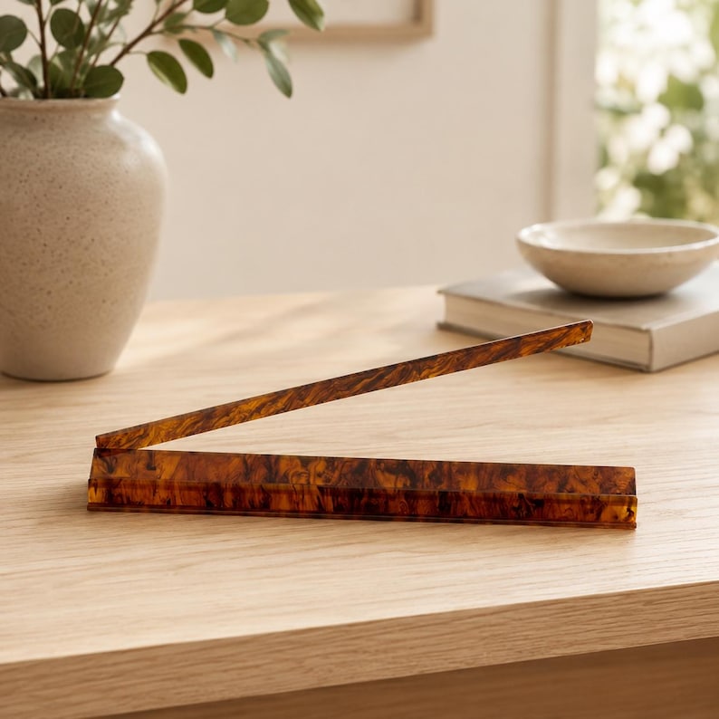 May include: A decorative, open book stand made of brown and amber-colored acrylic. The stand is on a light-colored wooden surface. A vase with greenery and a small bowl are in the background.