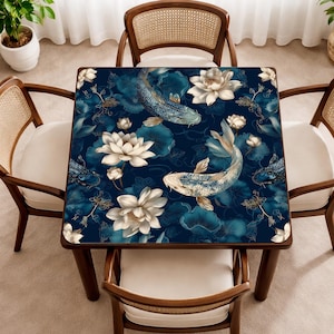 May include: Square dining table with a dark blue surface featuring a koi fish and lotus flower design. The table is surrounded by four wooden chairs with woven backs and white cushions. The table's design includes white and gold accents.