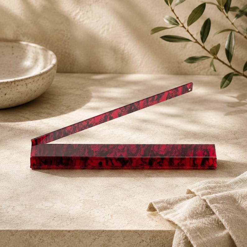 May include: A red and black marbled acrylic box with a hinged lid, open at an angle. The box is rectangular and sits on a light-colored surface. A beige towel is in the foreground.