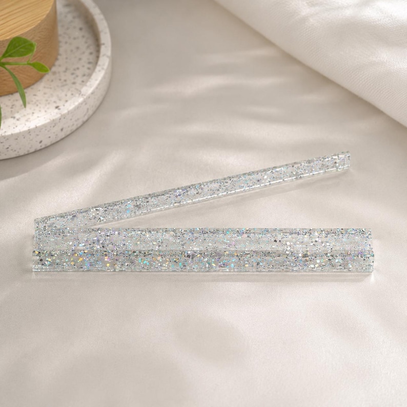 May include: Two clear, rectangular glitter rulers. The rulers are made of clear plastic and are filled with silver glitter. One ruler is open, and the other is closed. The rulers are on a white surface.