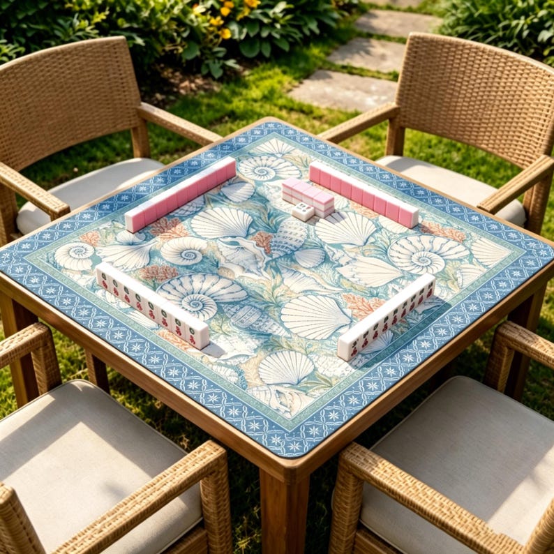 May include: A square game table featuring a blue and white seashell pattern on the surface, surrounded by four wicker chairs with light cushions. Four sets of white mahjong tiles are arranged in lines on the table, with one small die.