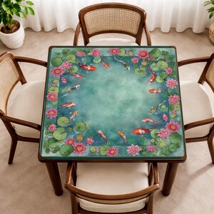 May include: Square wooden table with a teal surface featuring a koi pond design. The design includes pink lotus flowers, lily pads, and orange and white koi fish. The table is surrounded by four wooden chairs with cream-colored cushions.