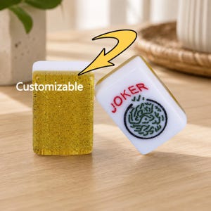 May include: Two Mahjong tiles, one gold and one white, are displayed on a wooden surface. The gold tile has the word "Customizable" printed on it. The white tile reads "JOKER" in red and features a green and black design.