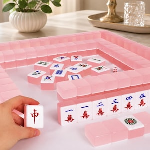 May include: A pink Mahjong set with translucent tiles and a matching tray. The tiles feature Chinese characters and symbols in red and blue. A hand holds a tile with a red character. A white die is also visible.
