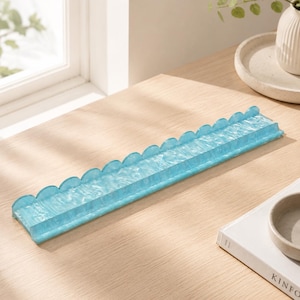 May include: A long, rectangular, light blue decorative tray with a scalloped edge. The tray is made of a translucent material and sits on a light wood surface. The tray is empty.