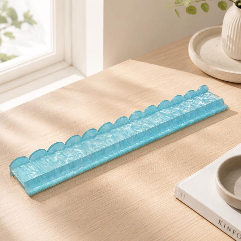May include: A long, rectangular, light blue decorative tray with a scalloped edge. The tray is made of a translucent material and sits on a light wood surface. The tray is empty.