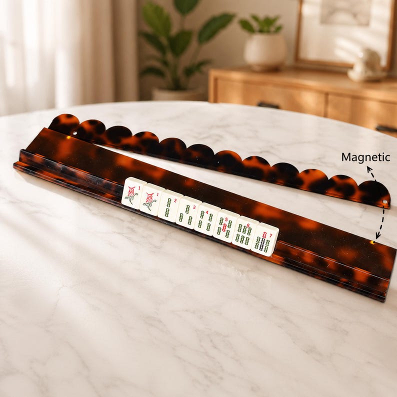 May include: A brown and amber-colored Mahjong tile holder with a matching magnetic tile rack. The holder is rectangular with a scalloped edge, and the tiles are white with red and green markings. The word "Magnetic" is written on the image.