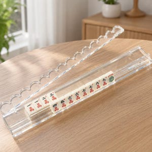 May include: A clear acrylic mahjong tile holder with a scalloped lid. The holder contains a row of white mahjong tiles with red and green Chinese characters. The holder is on a wooden table.