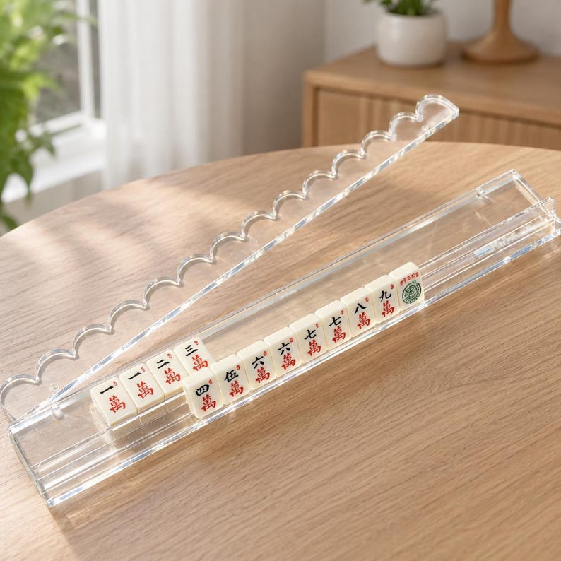 May include: A clear acrylic mahjong tile holder with a scalloped lid. The holder contains a row of white mahjong tiles with red and green Chinese characters. The holder is on a wooden table.