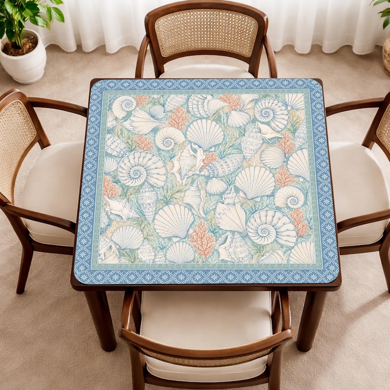 May include: A square table mat featuring a detailed pattern of various white seashells and orange coral branches on a light blue background, framed by a decorative blue border with a repeating white geometric star and diamond motif.