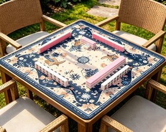 Puede incluir: Una mesa de mahjong cuadrada de madera con una parte superior estampada floral en crema y azul marino, rodeada de cuatro sillas de mimbre. La mesa está puesta con fichas de mahjong rosas y blancas, listas para jugar. La escena es al aire libre en un césped.