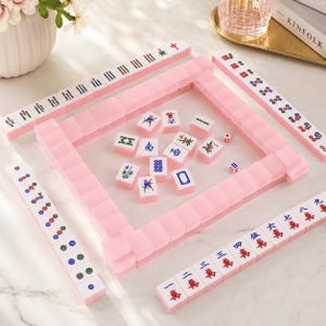 May include: A pink Mahjong set with tiles and racks arranged on a light surface. The tiles feature various symbols and characters in red, green, blue, and black. Two dice are also present, along with a vase of white flowers.