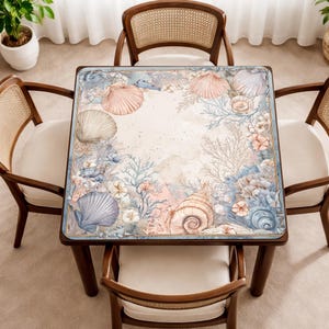 May include: A square table featuring a printed surface with a detailed marine-themed illustration. The design displays various seashells, coral formations, and sea plants in soft shades of blue, pink, and beige, arranged in a border pattern.