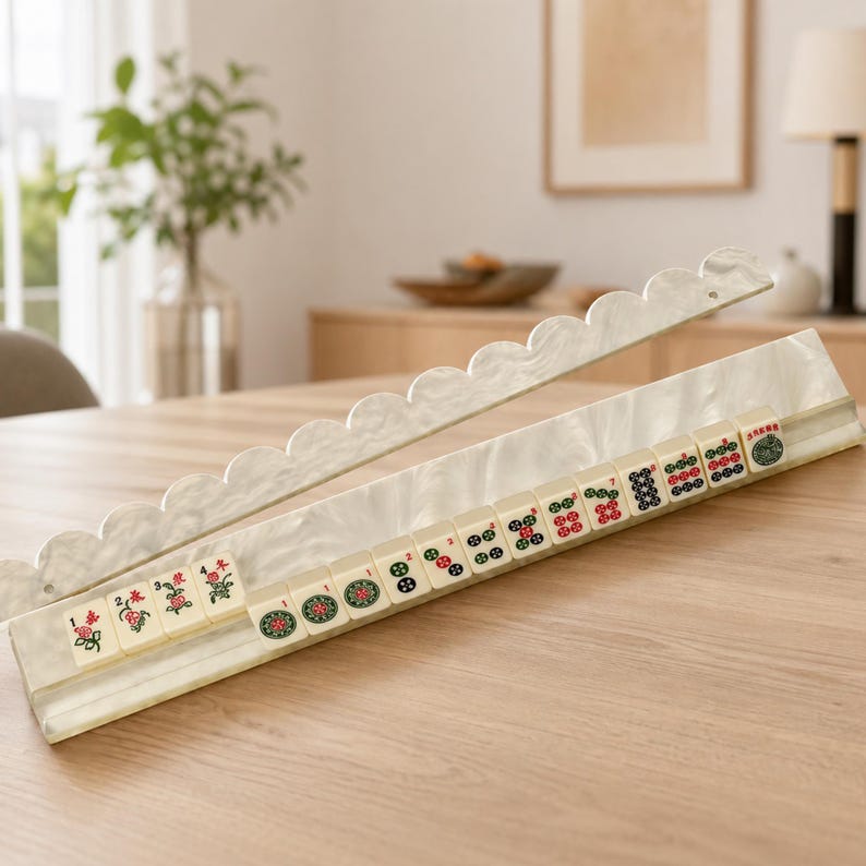 May include: A cream-colored Mahjong set with scalloped edges. The tiles are ivory with red, green, and black symbols. The set is displayed on a light wood table, showcasing the game's components and design.