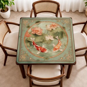 May include: A square table mat featuring a central circular illustration of two koi fish swimming among pink lotus flowers and green lily pads. The mat has a sage green background with ornate gold-toned filigree patterns in the corners.