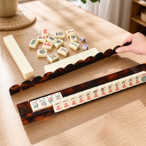 May include: A Mahjong set with ivory-colored tiles and a tortoise-shell patterned rack. The tiles have red and green symbols. Two dice are also present. The set is displayed on a light-colored wooden surface.