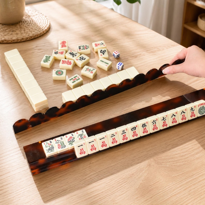 May include: A Mahjong set with ivory-colored tiles and a tortoise-shell patterned rack. The tiles have red and green symbols. Two dice are also present. The set is displayed on a light-colored wooden surface.