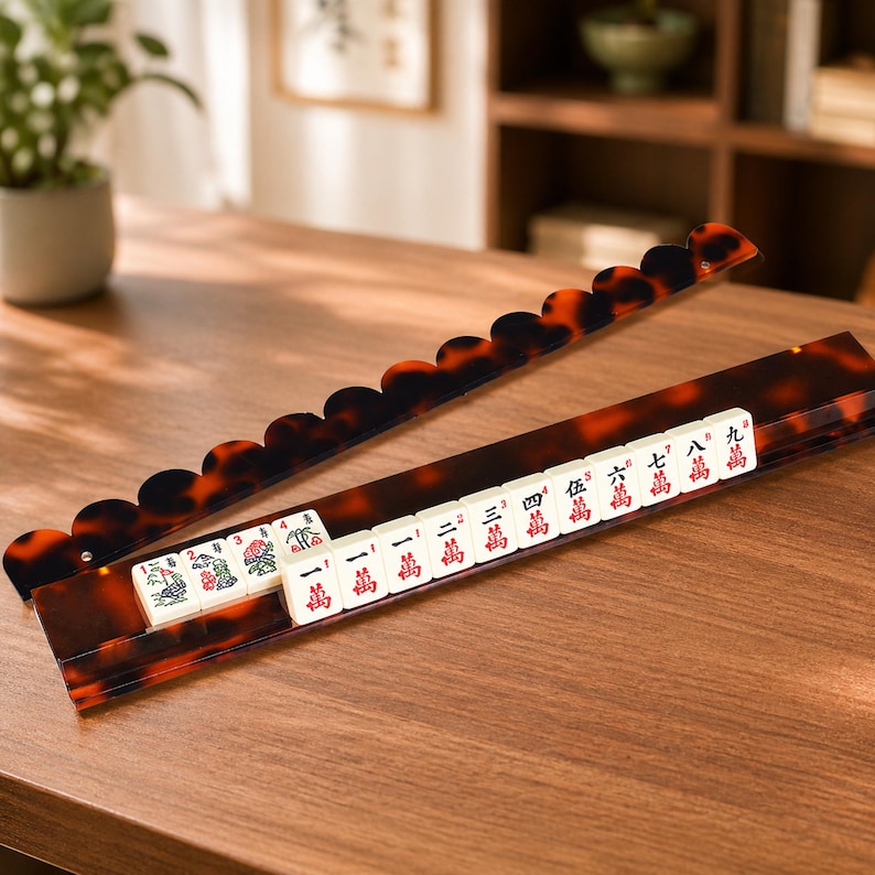 May include: A brown and black Mahjong set with a tortoise shell design. The set includes a rack and tiles with Chinese characters and symbols. The tiles are white with red and green markings. The rack has a scalloped edge.