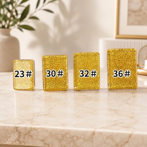 May include: Four gold-colored, rectangular objects with the numbers 23#, 30#, 32#, and 36# printed on them. The objects are arranged in a row on a light-colored surface. The background includes a vase with greenery and a framed picture.