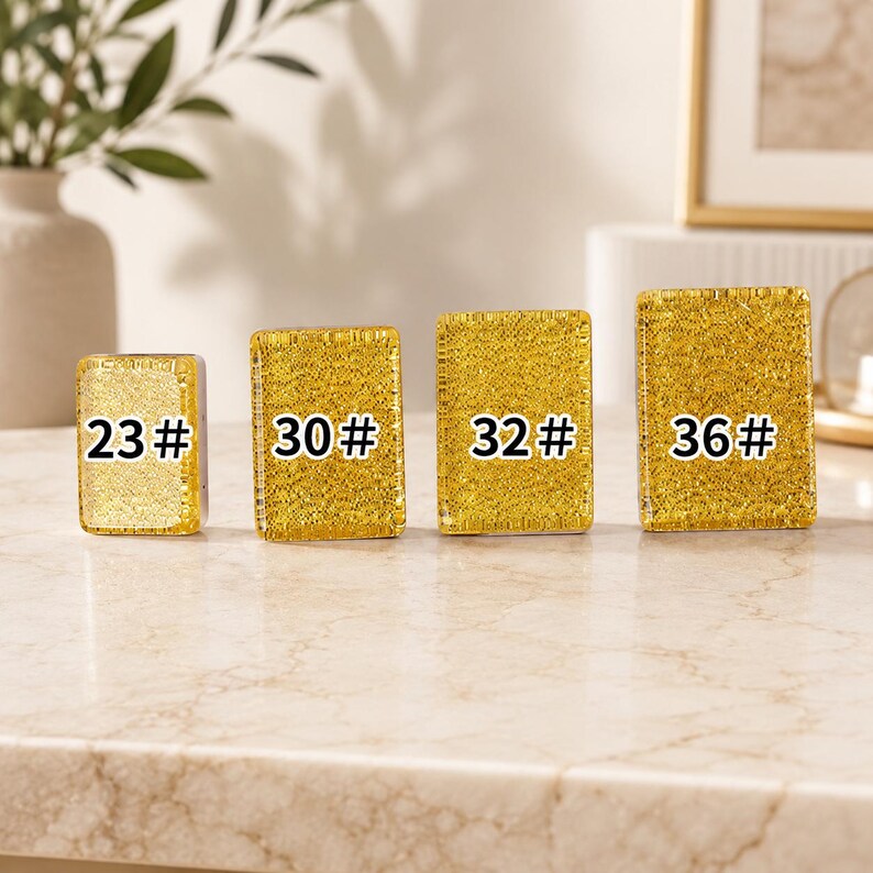 May include: Four gold-colored, rectangular objects with the numbers 23#, 30#, 32#, and 36# printed on them. The objects are arranged in a row on a light-colored surface. The background includes a vase with greenery and a framed picture.
