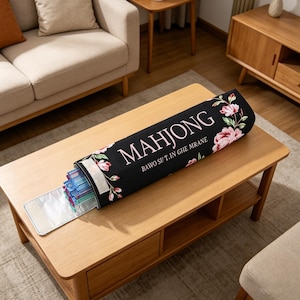 May include: A black Mahjong tile case with a floral design, open to reveal colorful tiles. The case has the word "MAHJONG" in pink letters and the text "BAWO SI/ T.IN GHE MRANE". The case is on a wooden coffee table.