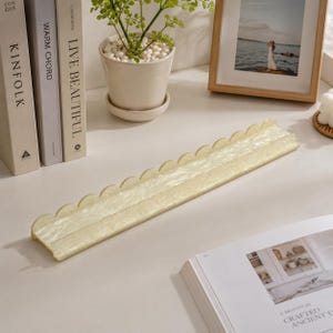 May include: A long, rectangular, scalloped-edge tray in a pale yellow hue. The tray is made of a translucent material and sits on a white surface. Decorative books, a framed photo, and a potted plant are in the background.