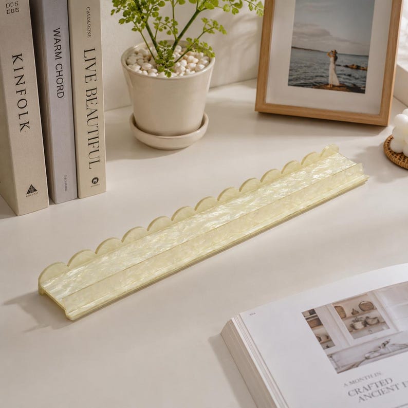 May include: A long, rectangular, scalloped-edge tray in a pale yellow hue. The tray is made of a translucent material and sits on a white surface. Decorative books, a framed photo, and a potted plant are in the background.