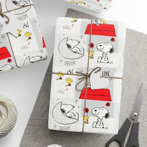May include: Gift-wrapped presents with Snoopy and Woodstock designs. The wrapping paper features Snoopy on his doghouse, sleeping on a crescent moon, and Woodstock singing. The gifts are tied with twine and adorned with tags that say "LOVE", "DREAM", and "SING".