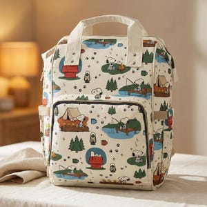 May include: Cream-coloured backpack with a repeating pattern of Snoopy camping scenes. The design includes Snoopy at a campfire, fishing, and in his doghouse. The backpack has a top handle, front pocket, and side pocket.
