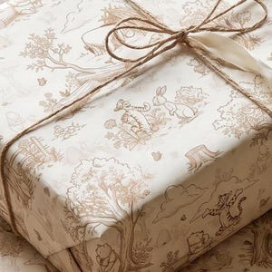 May include: A rectangular gift box wrapped in cream-colored paper featuring a repeating brown line-art illustration of Winnie the Pooh characters in a forest setting. The package is tied with a thin brown twine and a white ribbon bow.