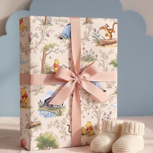 May include: A rectangular gift box wrapped in cream-colored paper featuring a repeating pattern of Winnie the Pooh characters, trees, and flowers. The box is tied with a dusty pink satin ribbon tied in a bow, accompanied by two small cream-colored knit booties.