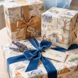 May include: Three square gift boxes wrapped in cream-colored paper featuring Winnie the Pooh illustrations. The boxes are adorned with gold, cream, and blue velvet ribbons, a bee charm, a sprig of lavender, and tags labeled Pooh and For You.