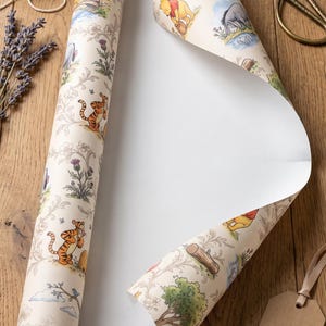 May include: A roll of cream-colored wrapping paper featuring a repeating pattern of Winnie the Pooh characters including Pooh, Tigger, and Eeyore, interspersed with floral and woodland illustrations. The paper has a smooth, matte finish.