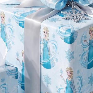 May include: A square gift box wrapped in light blue paper featuring a repeating pattern of a blonde character in a blue dress surrounded by snowflakes. The package is adorned with a wide silver ribbon and a large, glittery silver snowflake ornament.