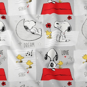 Puede incluir: Tela con un patrón repetitivo de Snoopy y Woodstock en varias poses. Snoopy se muestra en su caseta, en una luna creciente y con Woodstock. También se incluyen las palabras "LOVE", "DREAM" y "SING".
