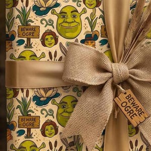 May include: Wrapping paper featuring a repeating pattern of Shrek characters, including Shrek, Fiona, Donkey, and Puss in Boots, alongside cattails, water lilies, and wooden signs that read BEWARE OGRE, tied with a tan burlap and satin ribbon.